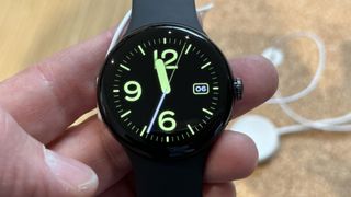 Google Pixel Watch being held in a person's hand with a green analogue clock face on the display