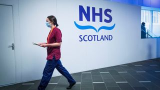 NHS Scotland sign pictured at a vaccination center with nurse in scrubs walking by.