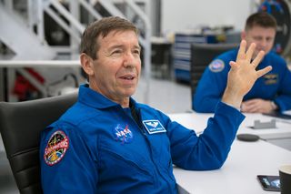 a man wearing a blue jumpsuit sits in a white room with black furniture. his elbow rests on a table.