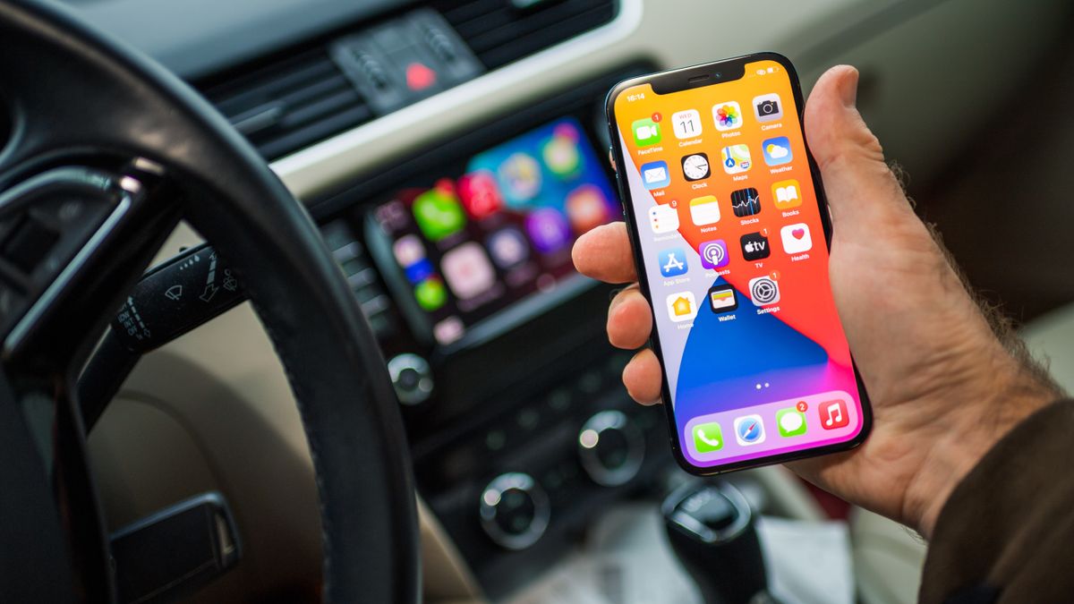 Man&#039;s hand holding iPhone in car with CarPlay screen behind