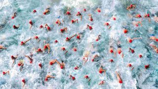 Top-down image of tens of swimmers with red swimming caps creating loads of splashes in the water 