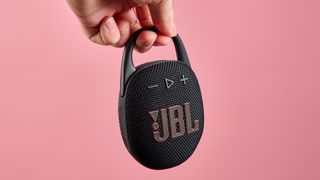 The JBL Clip 5 in black is pictured being held by the carabiner against a pink background,