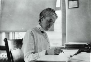 Henrietta Swan Leavitt sitting at a desk holding a pen and looking at books.