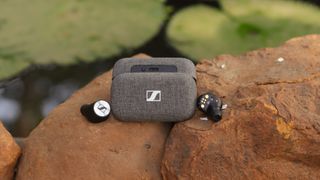 Bästa in-ear-hörlurar med brusreducering: Ett par Sennheiser Momentum True Wireless 2 ligger utomhus på en sten bredvid sitt tillhörande grå laddningsfodral.