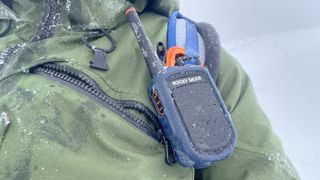 Close up of a Rocky Talkie Mountain Radio walkie-talkie attached to a blue backpack strap against a green ski jacket covered in ice with snow in the distance