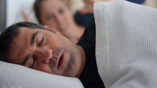 A man with sleep apnea snores while partner looks exasperated in the background