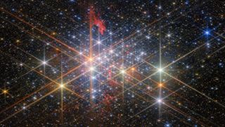 A dense cluster of bright stars, each with six large and two small diffraction spikes, due to the telescope’s optics. They have a variety of sizes depending on their brightness and distance from us in the cluster, and different colors reflecting different types of star. Patches of billowing red gas can be seen in and around the cluster, lit up by the stars. Small stars in the cluster blend into a background of distant stars and galaxies on black.