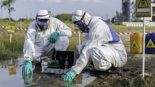 Two people in hazmat suits collect water samples near a waste processing plant