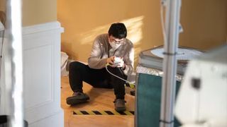 scientist wearing a face mask and glasses sits on the floor of a house while taking samples from a small device in his hands