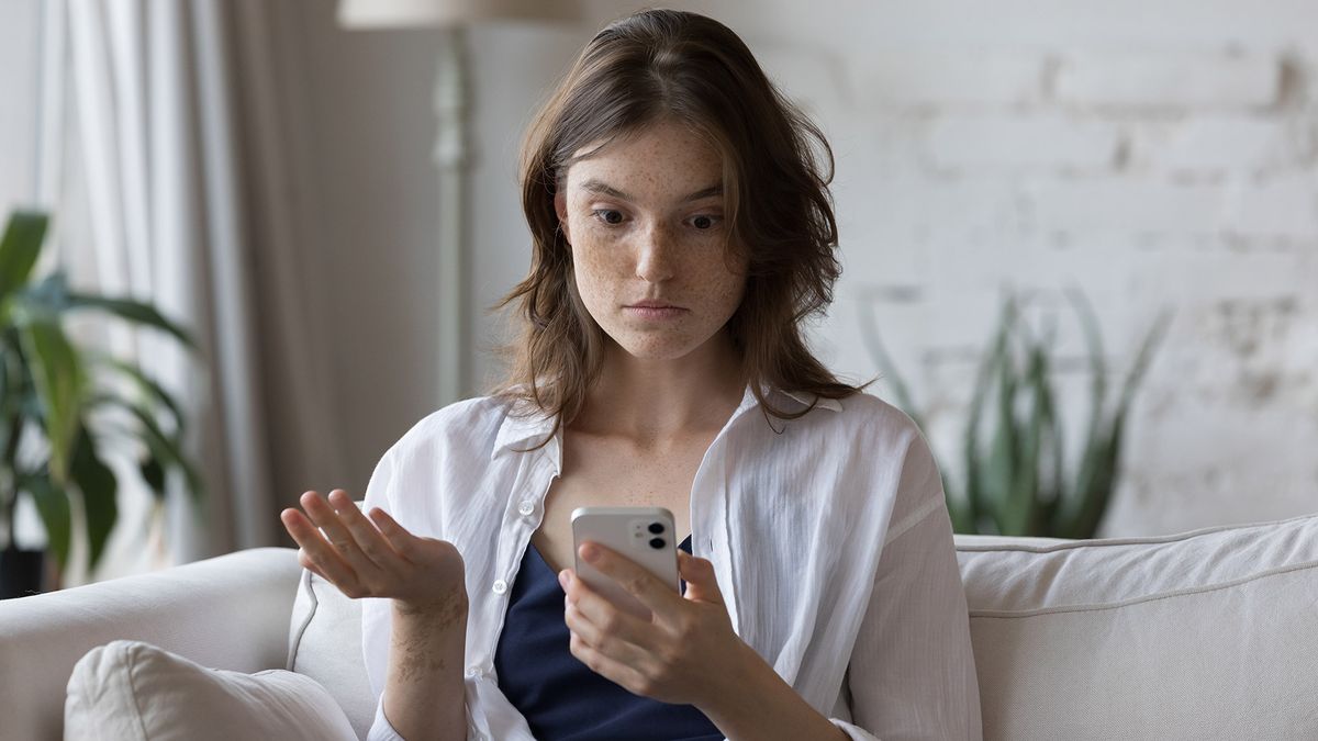 A woman sitting in a living room stares at a phone in confusion