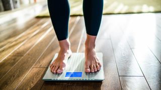 A woman&#039;s feet are shown standing on weighing scales that are placed on a wooden floor. She is wearing black leggings and nothing on her feet.