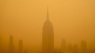 photo shows the empire state building as viewed from a nearby skyscraper; the building and those nearby are surrounded by a dense cloud of yellow-orange smog made up of wildfire smoke