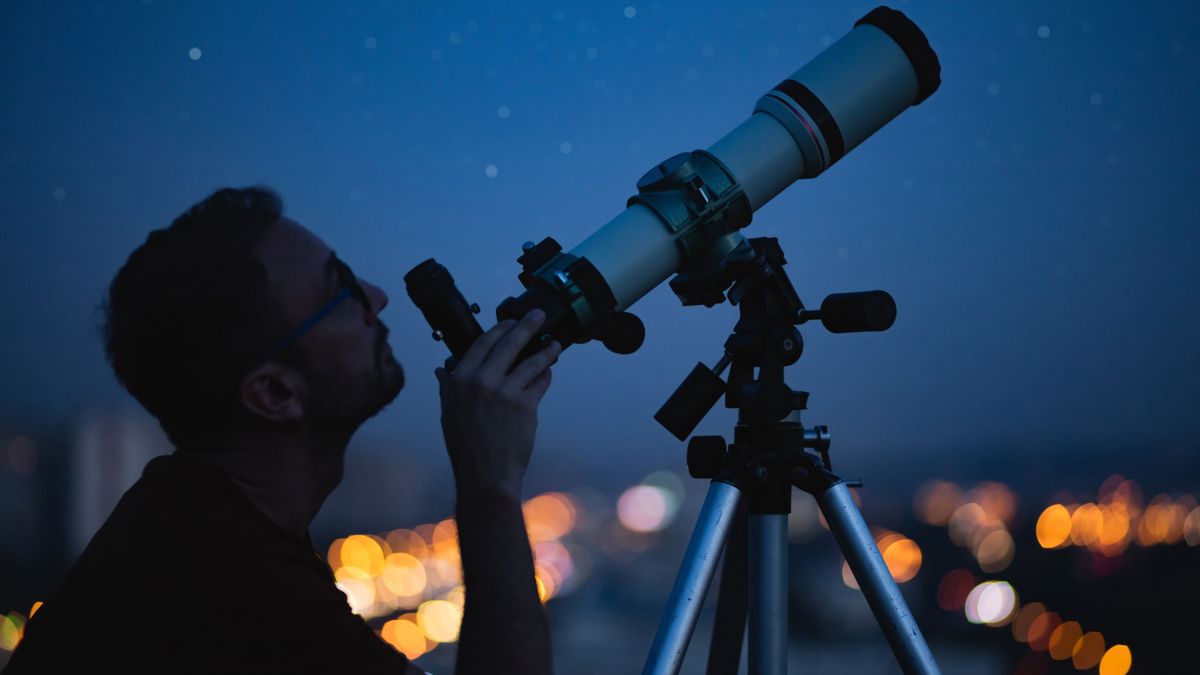 Image shows man using telescope with city lights in background