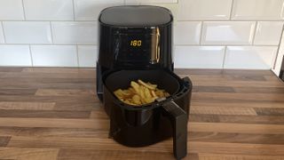 En svart Philips Essential Airfryer står på en träfärgad köksbänk med behållaren fylld med pommes frites, redo att tillagas.