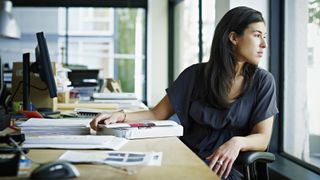 A business woman sitting at her desk but turned away from it looking wistfully out a window