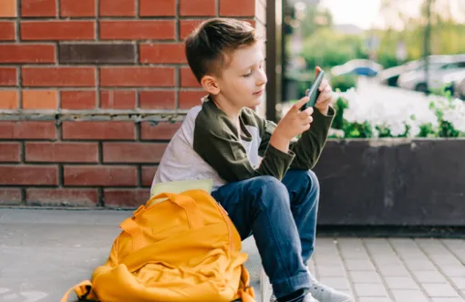 A child sitting on the stoop of a building, browsing their phone, with a yellow backpack beside them