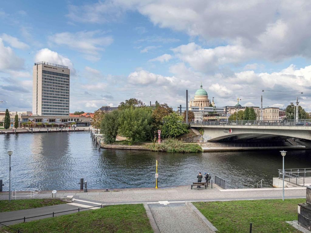 una persona sentada en un banco junto a un río con un puente en Mercure Hotel Potsdam City en Potsdam