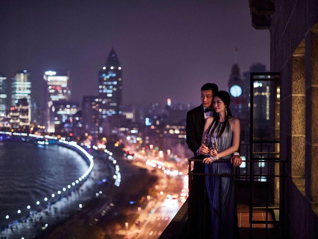 um homem e uma mulher de pé numa varanda à noite em Fairmont Peace Hotel on the Bund em Xangai