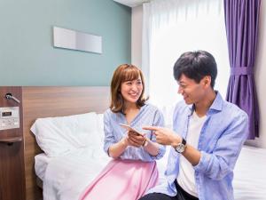 a man and a woman sitting on a bed looking at a cell phone at Comfort Hotel Kitakami in Kitakami