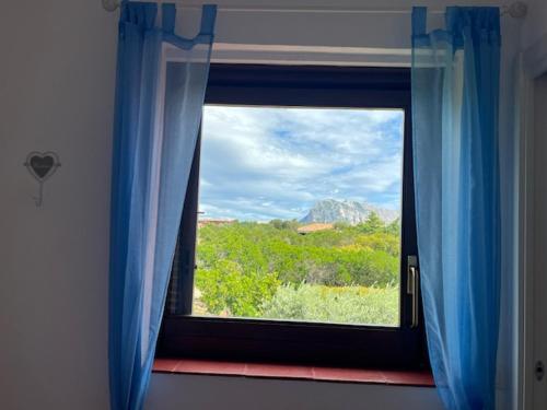 ein Fenster mit blauen Vorhängen und Bergblick in der Unterkunft Porto Coda Cavallo Salina Bamba in Capo Coda Cavallo