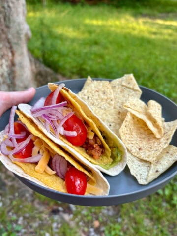 Camp tacos on plate with crunchy shell inside flour tortilla and filled with meat, veggies and cheese.