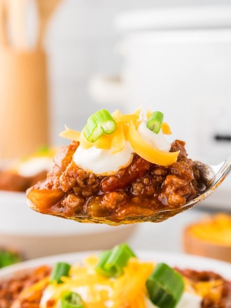 A spoonful of chili with cheese and sour cream in front of a slow cooker.