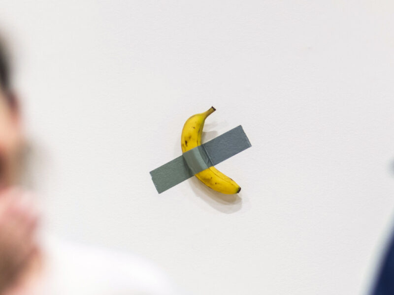 People react to the artist Maurizio Cattelan's piece of art "Comedian" during an auction preview at Sotheby's in New York. In the foreground, two out-of-focus people face away from the piece, laughing. In the background, in focus, is a yellow banana duct taped to a wall.