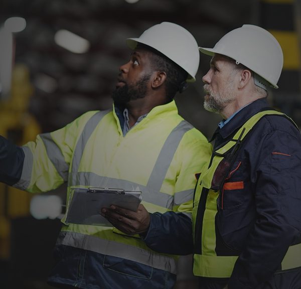 Two men in hard hats and safety gear standing with a clipboard pointing at an object off screen