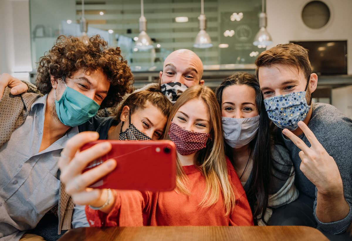 friends at home wearing masks posing for selfie: Shutterstock: Group of friends wearing protection mask at the restaurant - Young happy people celebrating taking a selfie with smartphone - People, technology and new normal lifestyle concept.