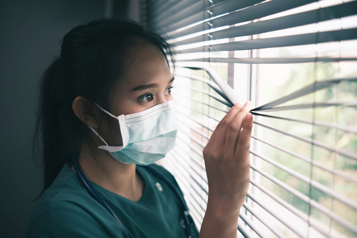 Woman wearing a mask and peeking out of the blinds looking scared: Asian female healthcare worker doctor nurse surgeon wearing protective surgical mask corona covid-19 virus prevention, feeling worried anxious anxiety traumatized scared, medical stress at hospital Shutterstock