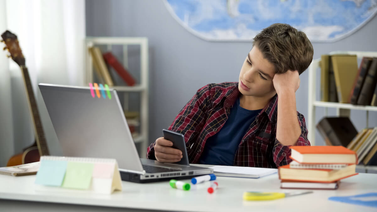 Boy looking at phone procrastinating homework: Shutterstock. Bored guy scrolling apps on phone, distracted from homework, procrastination