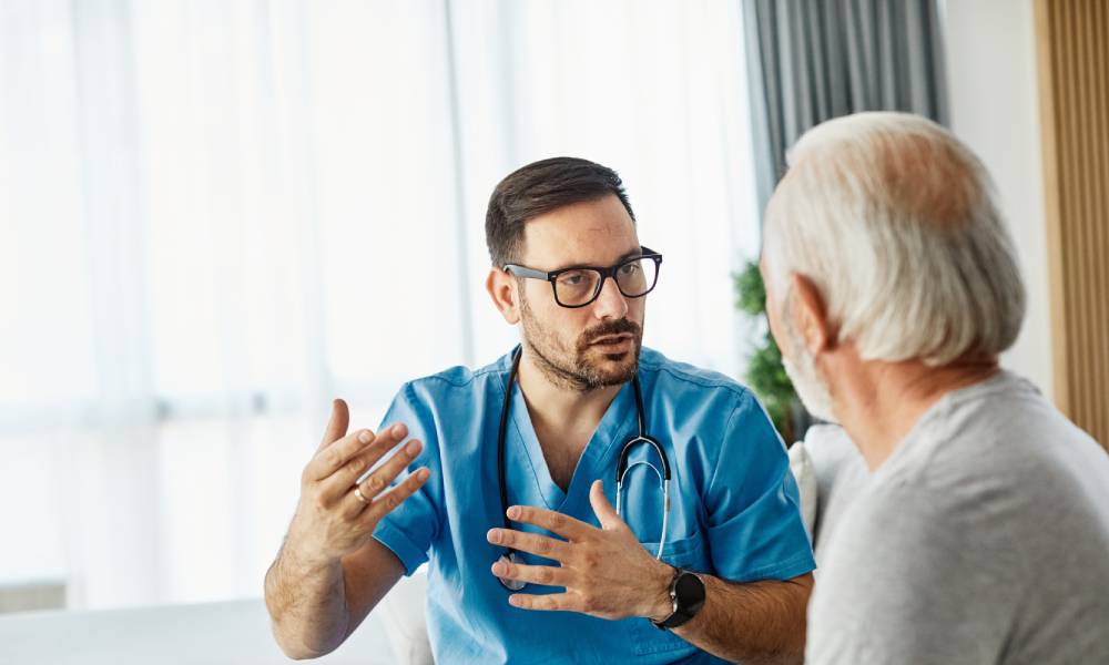 A medical professional in standard blue scrubs gesturing as they explain something to a senior patient.