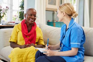 Black patient in yellow dress and red scarf smiling and sat on a sofa next to a community nurse. The nurse is white with blonde hair and is wearing a blue uniform and holding a notebook and pen.