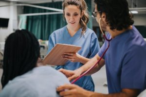 Picture of student nurses on a ward. One white female nurse is stood over a patient holding a clipboard. The other student nurse is using a stethoscope on the patient's back.