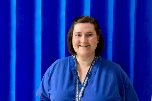 Headshot of Sarah Huggett. She is wearing a blue nurse uniform and is stood in front of a blue curtain, smiling.