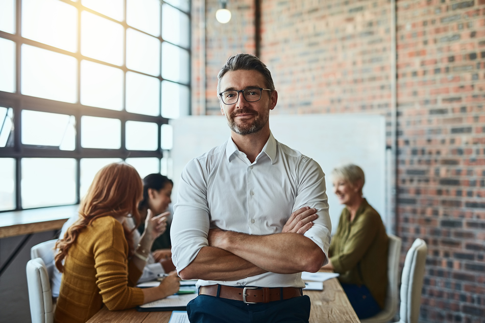 smiling man stands with arms crossed with office meeting happening behind him