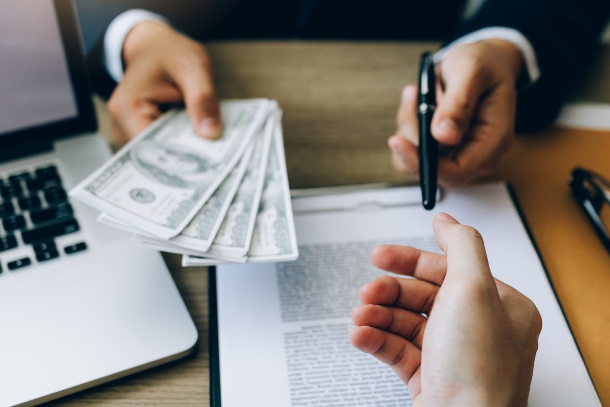 a person hands cash and a pen to a person across a desk holding a laptop and a loan agreement