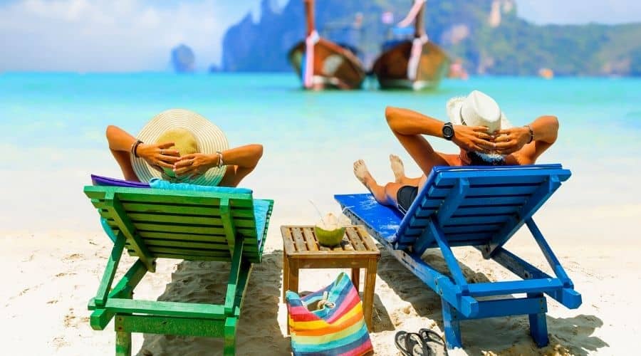 a man and woman wearing swimsuits and sunhats relax on lounge chairs at the beach