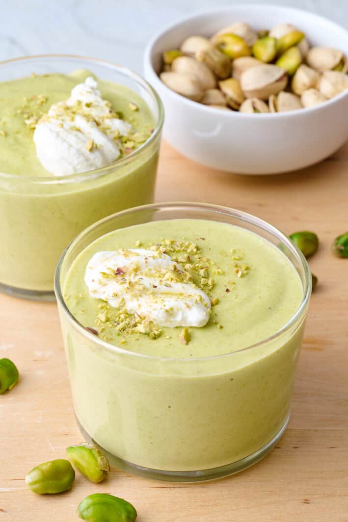 Creamy pistachio pudding in two serving glasses with small dish of pistachios nearby.