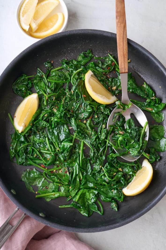 Sauteed spinach in skillet with a few lemon wedges and serving spoon dipped inside pan.