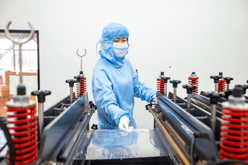 A worker wearing a blue body suit, face mask, and disposable gloves reaches out one hand to grasp a sheet of clear plastic packaging material from a table of clamps.