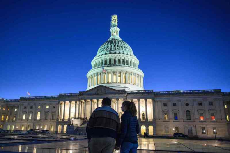 A couple stands with their back to the camera across a plaza from the dome of the U.S. Capitol building, which rises up against a bright blue dusk sky.