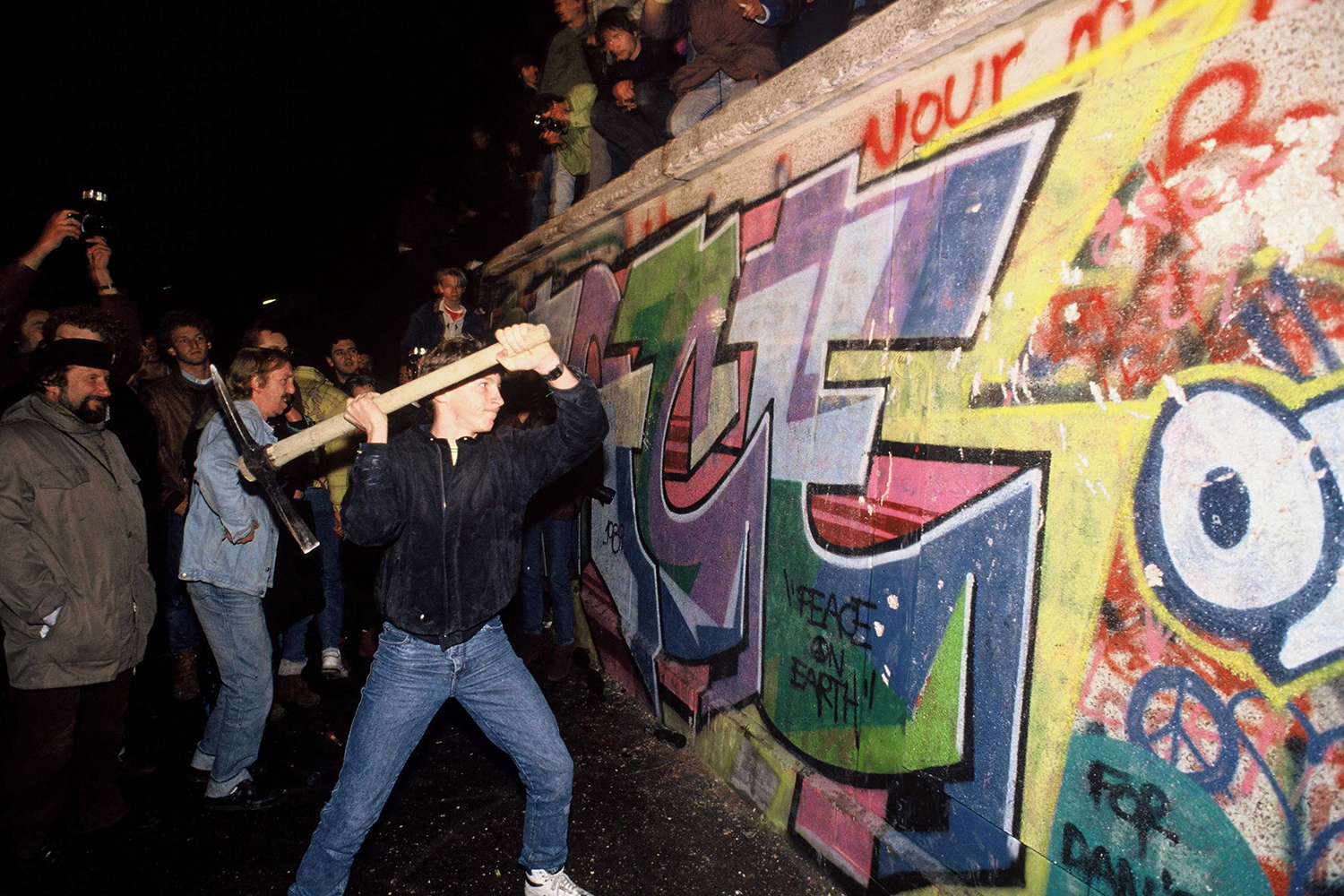 A man wearing jeans and a dark jacket lifts a pick ax to slam into a graffiti-covered section of the the Berlin Wall. Other protesters are seen behind him.