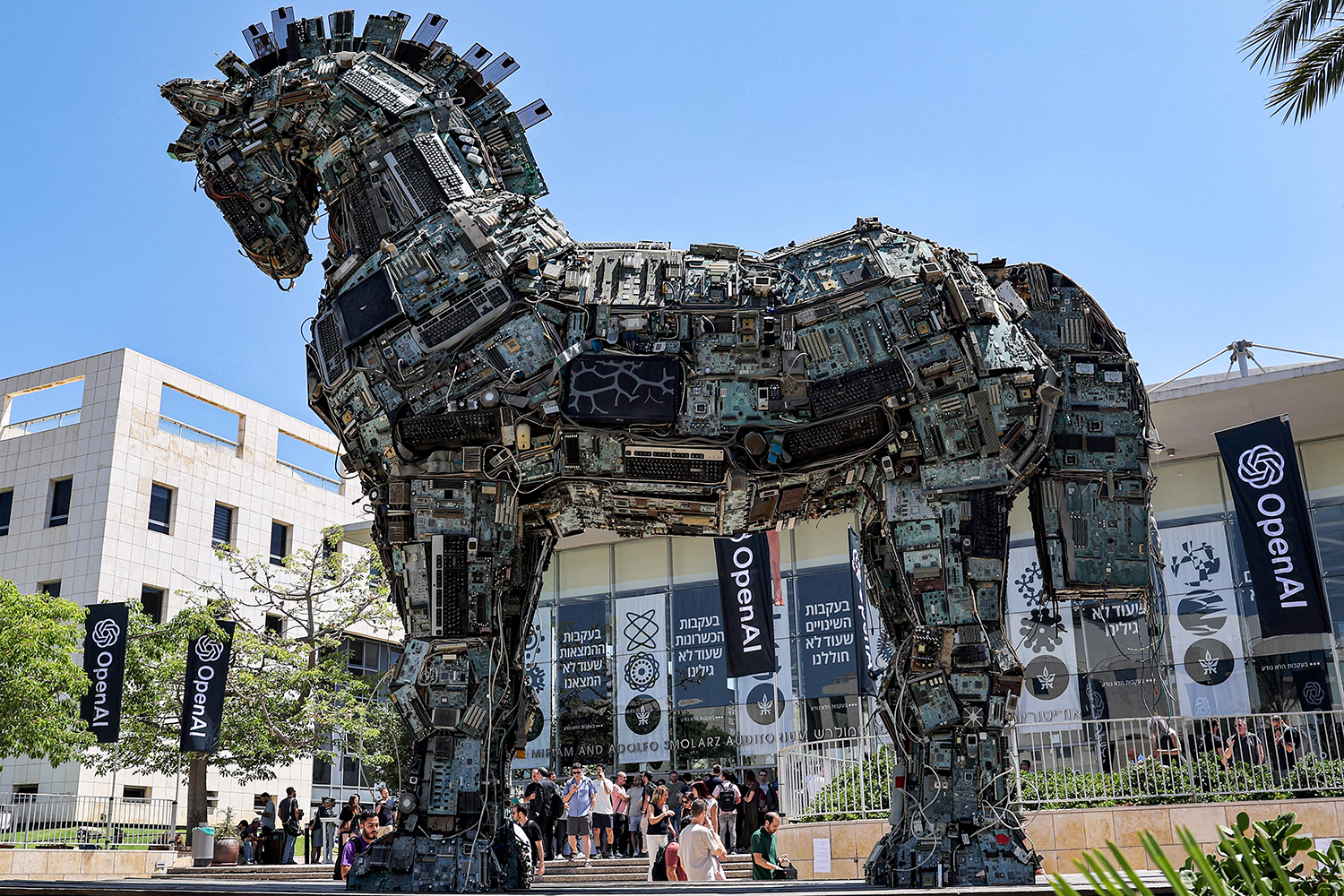 A giant trojan horse installation made up of electric components stands on a street. Behind it is a university building draped with OpenAI banners.