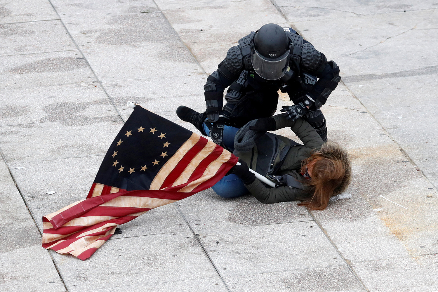 A police officer in riot gear pins down a protester holding a 13-star U.S. flag on the marble surface outside the Capitol.