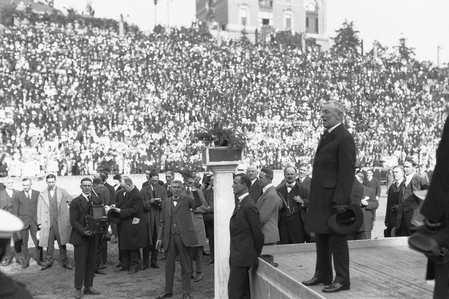 U.S. President Woodrow Wilson wearing a dark suit and holding a hat stands on a stage in front of a giant crowd in the stands. In the foreground, men in suits, one with a camera, cluster in front of him.