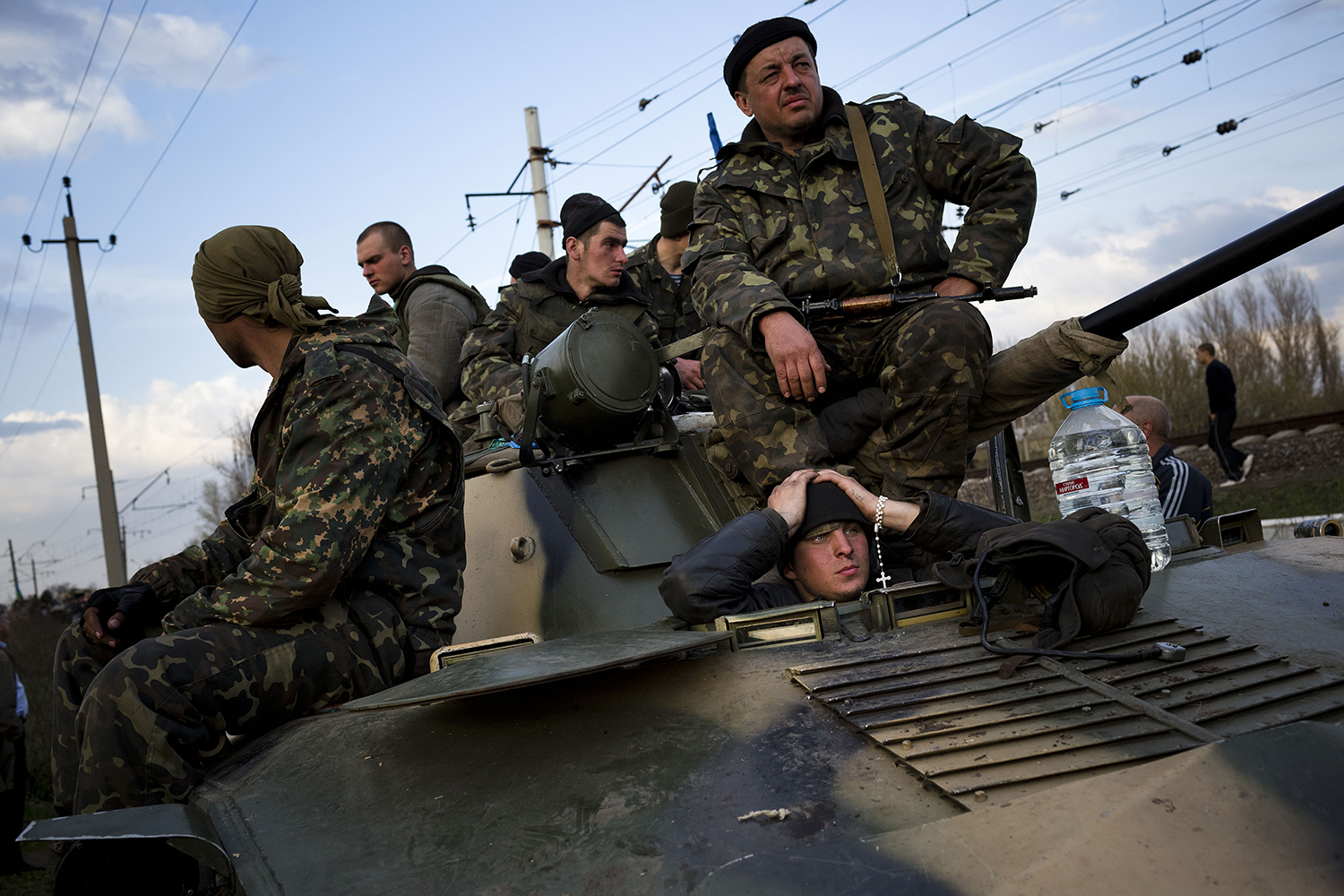 Five Ukrainian soldiers wearing camouflage are seen from below as they sit atop a tank. Another young soldier sticks his head out of a hatch from inside the tank and puts his hands to his head as he watches with a rosary wrapped around his wrist. Power lines are loom overhead against a partly cloudy blue sky.