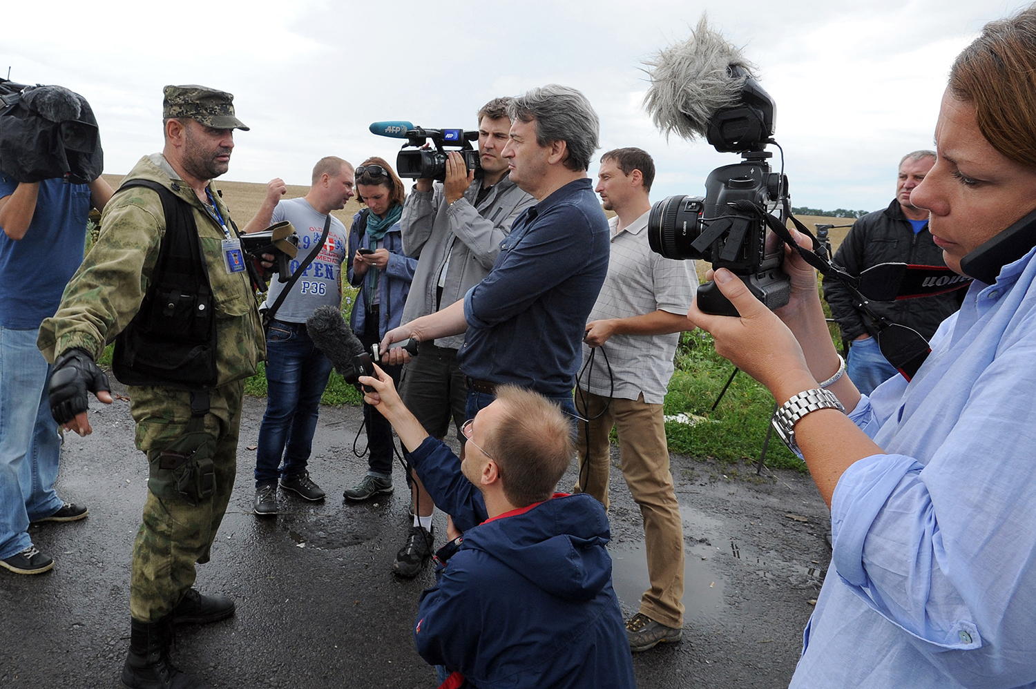 Journalists holding cameras and microphones cluster around an armed militant in olive green fatigues and a vest as he tries to block their path on a muddy road.