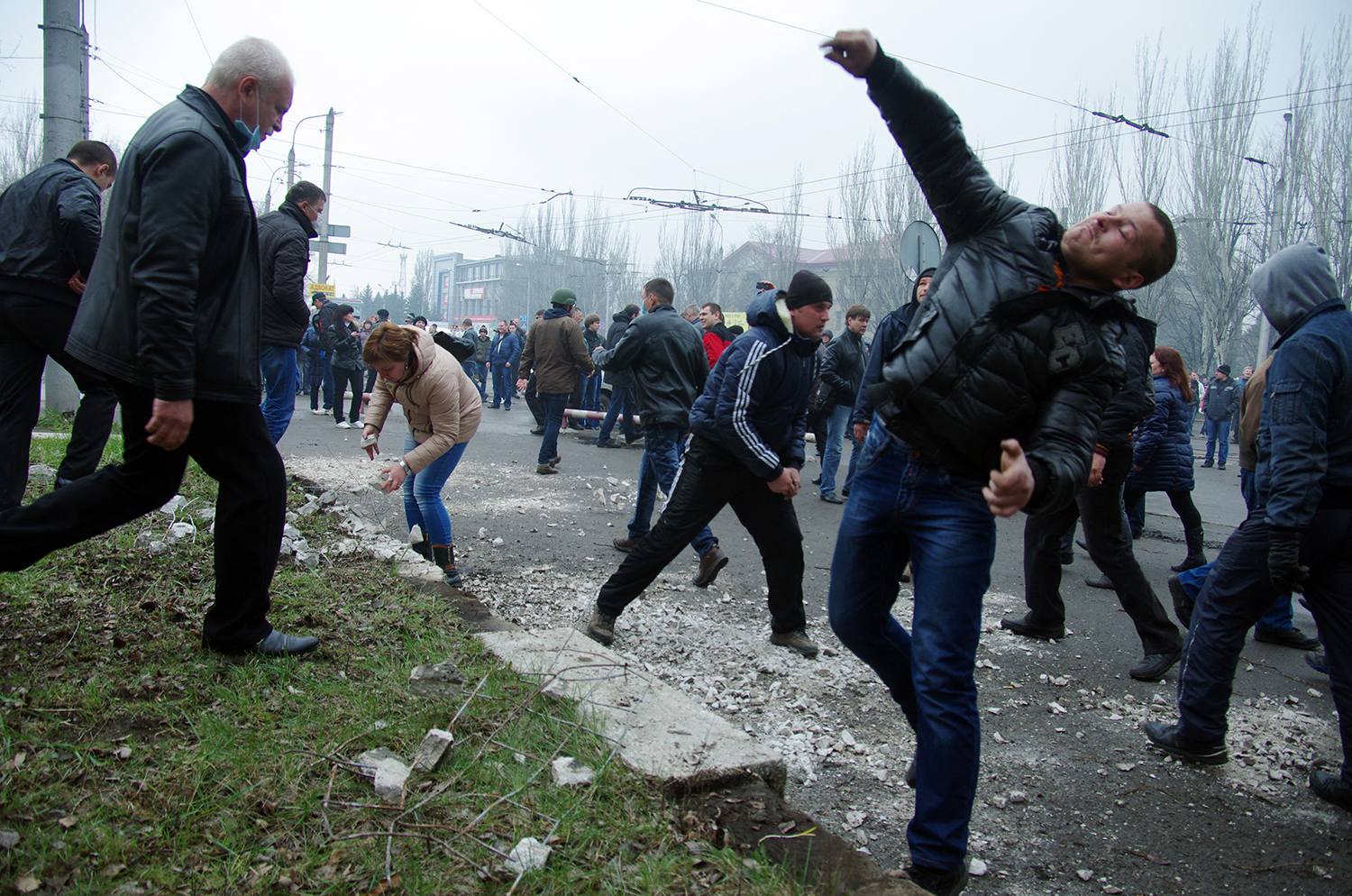 A man grimaces and arches his body as he throws a stone. Behind him, dozens of people mill about a road at a chaotic scene at a protest, with a few people in the foreground running or crouching to gather more crumbled pieces of stone.