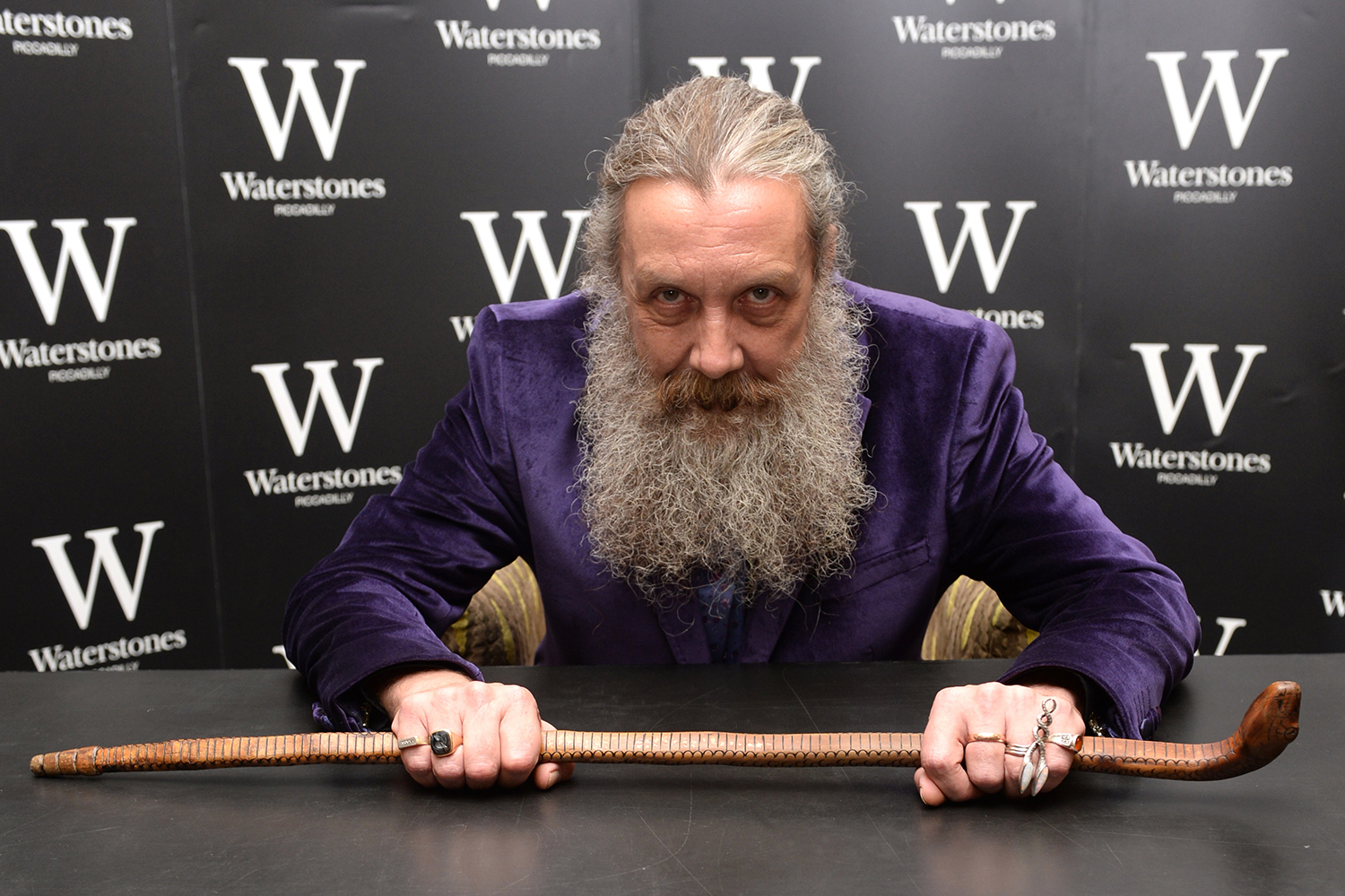 Alan Moore sits at a table while holding a cane in the shape of a snake.